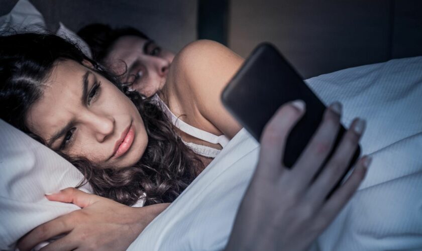 Paar liegt im Bett, sie sieht auf ihr Handy, er blickt misstrauisch hinüber zu ihr