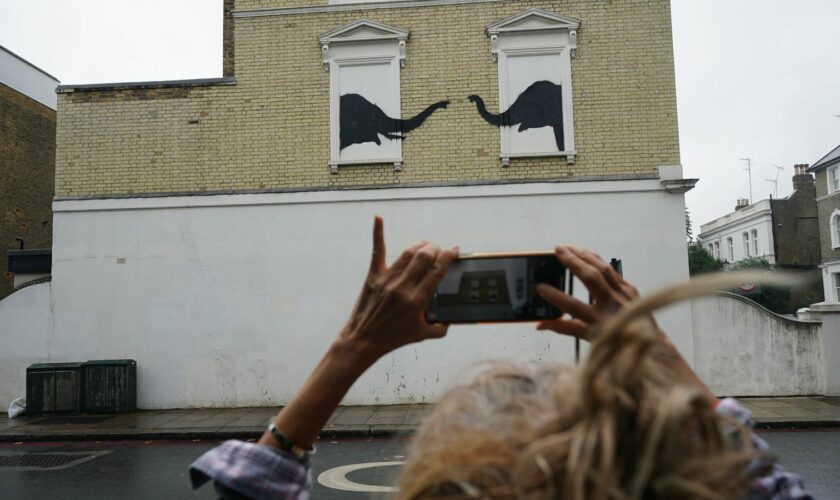 A woman stops to photograph a new artwork depicting two elephants poking their heads out of blocked out windows unveiled by Banksy on the side of a building at the junction of Edith Grove and Edith Terrace, in Chelsea, south west London. Picture date: Tuesday August 6, 2024.