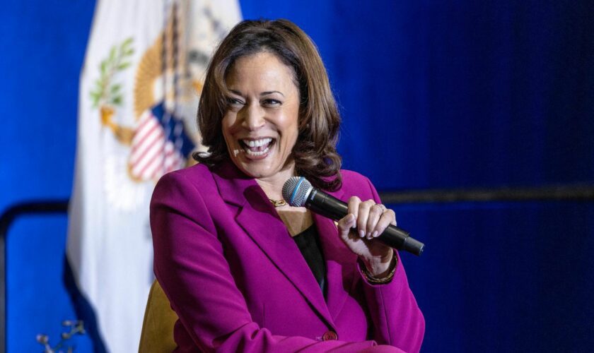 Kamala Harris sitzt auf einer Bühne und lacht, während sie mit links ein Mikro hält