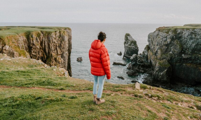 Eine Frau steht an einer Steilküste und trägt eine rote Steppjacke.