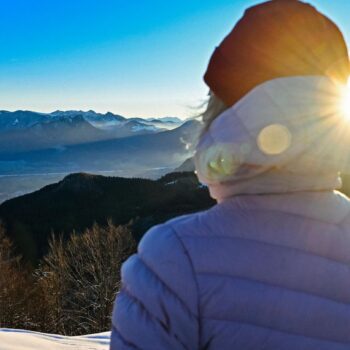 Eine Frau in dicker Jacke schaut bei Sonnenschein über Berge mit Schnee