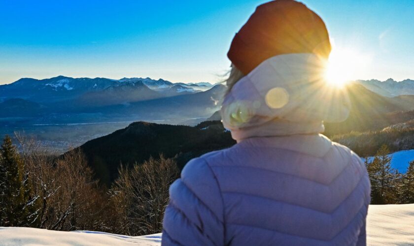 Eine Frau in dicker Jacke schaut bei Sonnenschein über Berge mit Schnee